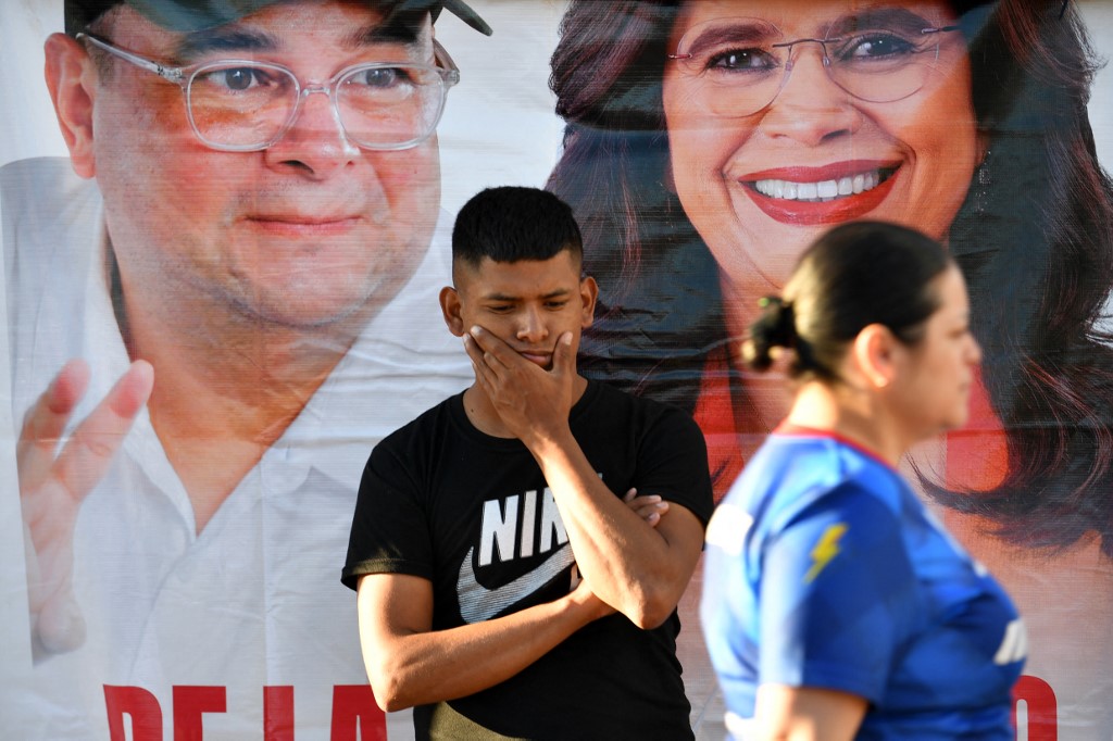 Um homem permanece em frente a um banner do prefeito de Tegucigalpa, Jorge Aldana, que busca a reeleição, e da candidata presidencial pelo partido Libertad y Refundación (Libre), Rixi Moncada, nas imediações das instalações do Instituto Nacional de Formação Profissional (INFOP), onde estão sendo contadas as cédulas das eleições gerais hondurenhas, em Tegucigalpa, em 10 de dezembro de 2025.