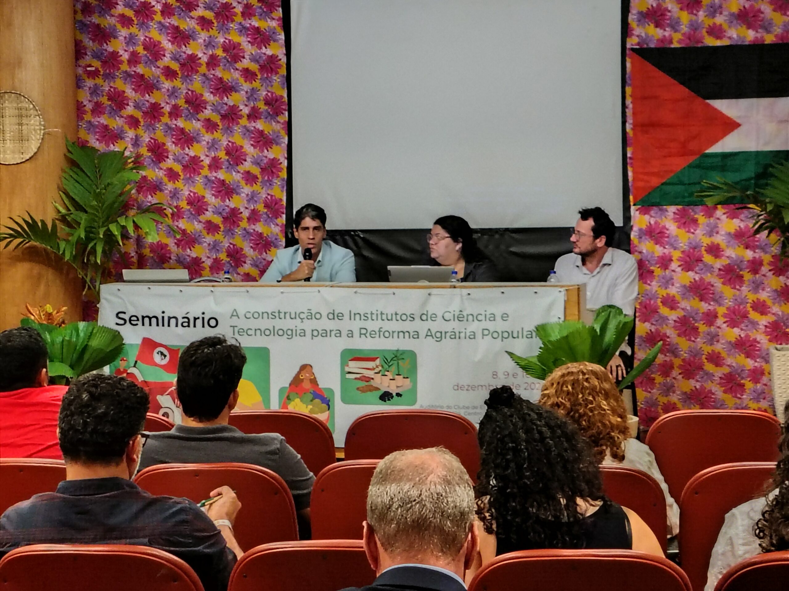 Joaquim Vasconcelos, representante do BNDES está com o microfone ao lado de Débora Nunes, dirigente nacional do MST e Daniel Mancio, da direção nacional. Eles estão em um auditório durante o Seminário de construção da estratégia de inovação das cooperativas e instituições científicas e tecnológicas da reforma agrária popular