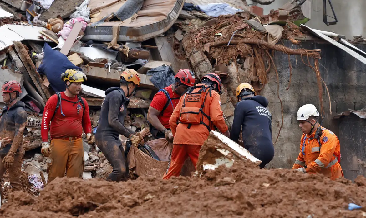 Soldados do Corpo de Bombeiros e voluntários fazem busca e resgate de pessoas em escombros de casas soterradas por lama após fortes chuvas.