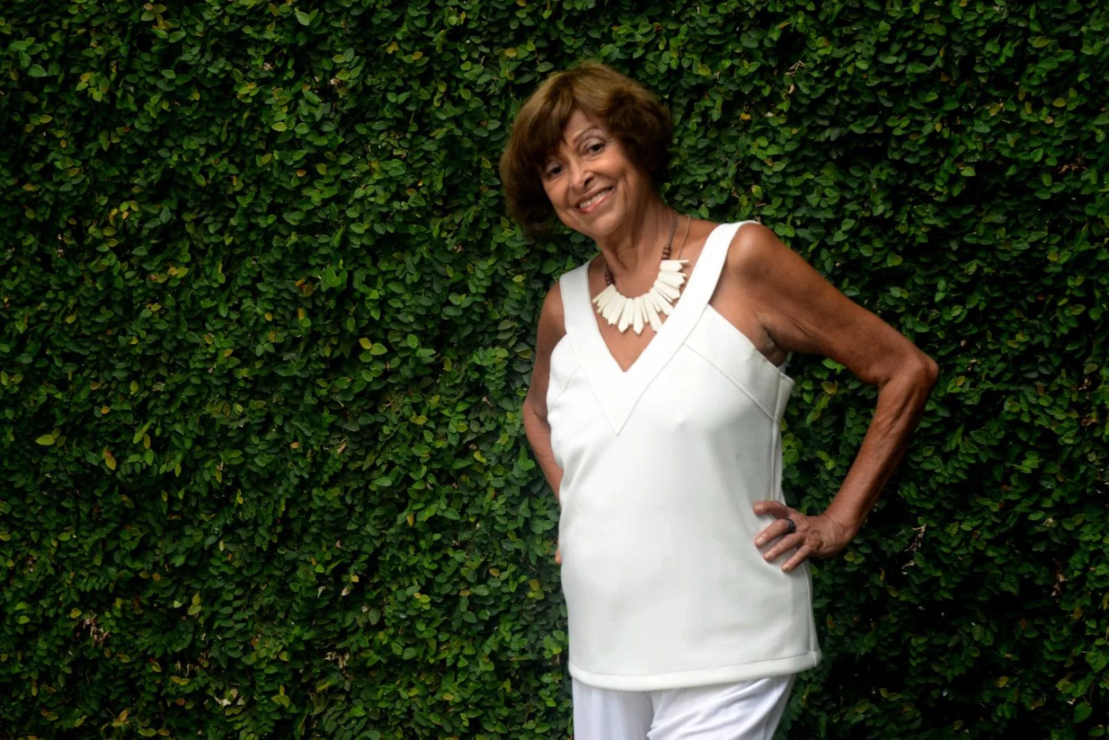 A cineasta Tete Moraes posa para foto vestida com a uma blusa longa, branca de alça em decote V e uma calça também branca. Ela tem cabelos curtos em formato channel e usa um colar de pedras retangulares igualmente brancos