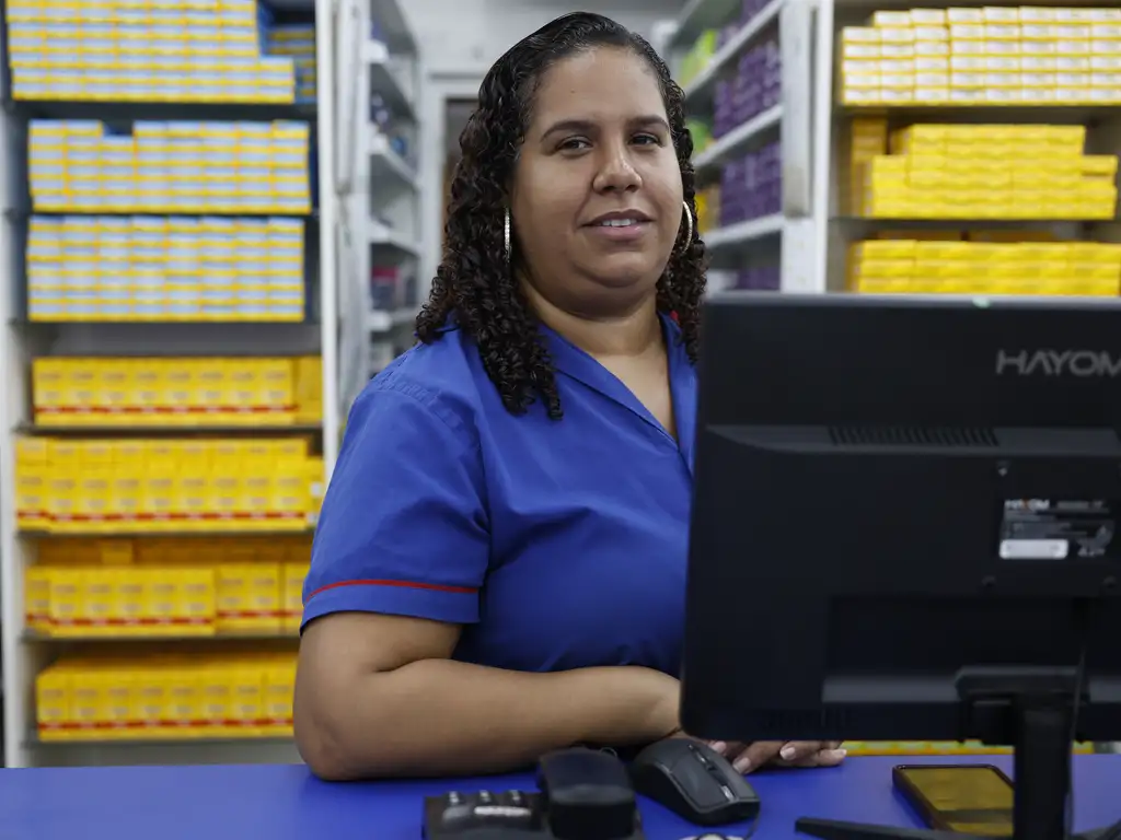 Mulher negra, estatura média e cabelos na altura do ombro é registrada em seu posto de trabalho. Balconista de farmácia, ao fundo está uma estante com vários remédios e no balcão há um computador na sua frente