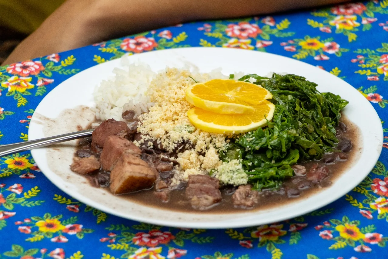 Prato de feijoada com carnes, arroz, uma rodela de laranja e couve sobre mesa com pano colorido