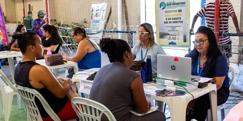 Em um ginásio poliesportivo, mesas de cadeiras de plástico são dispostas para atender a população. Na foto, há duas atendentes, cada uma com seu laptop, atendendo duas mulheres