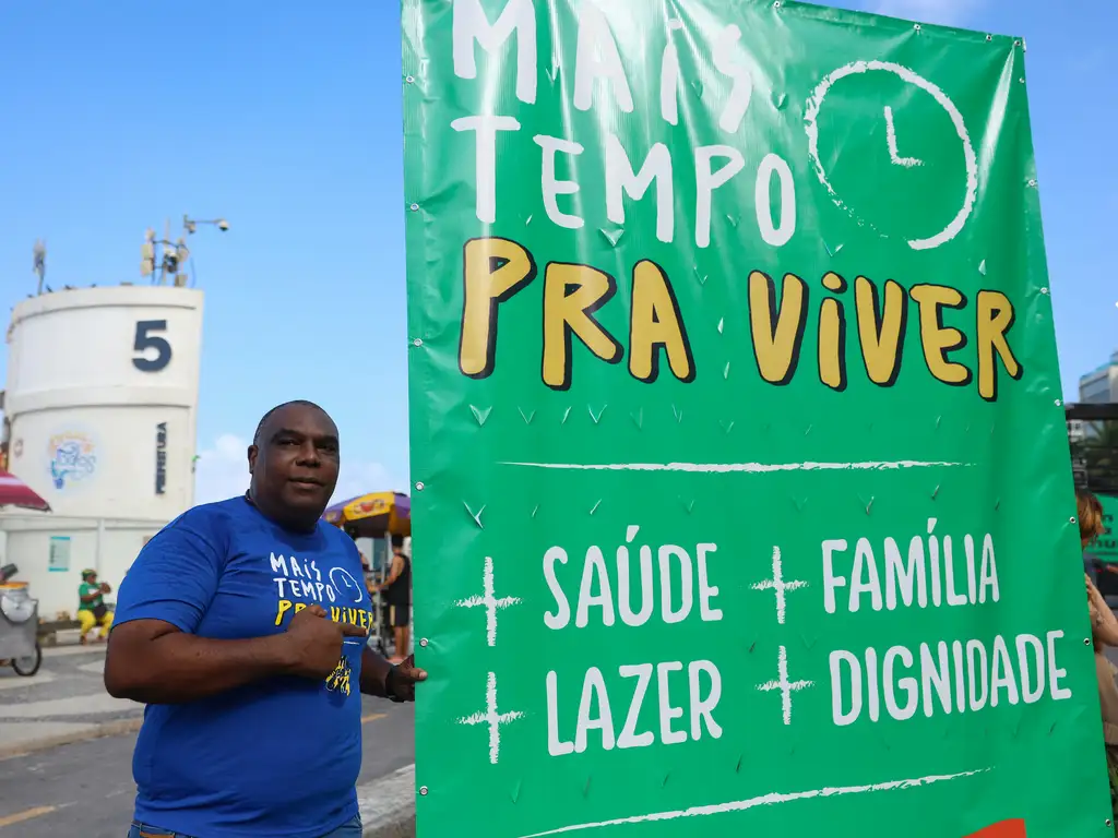 Em manifestação em Copacana, homem negro posa ao lado de uma placa de fundo verde clara em que está escrito 'mais tempo para viver' em destaque e na sequência estão mais saúde, mais lazer, mais família, mais dignidade