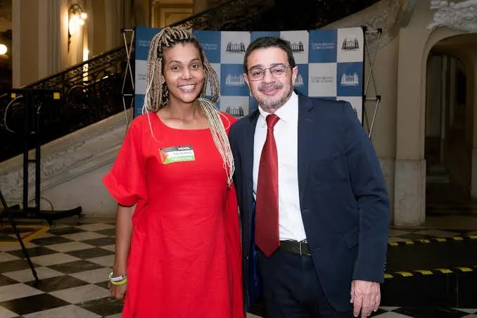 Dani Balbi, com vestido longo vermelho, posa ao lado de Elias Jabbour, de terno escuro, blusa social branca e gravata vermelha